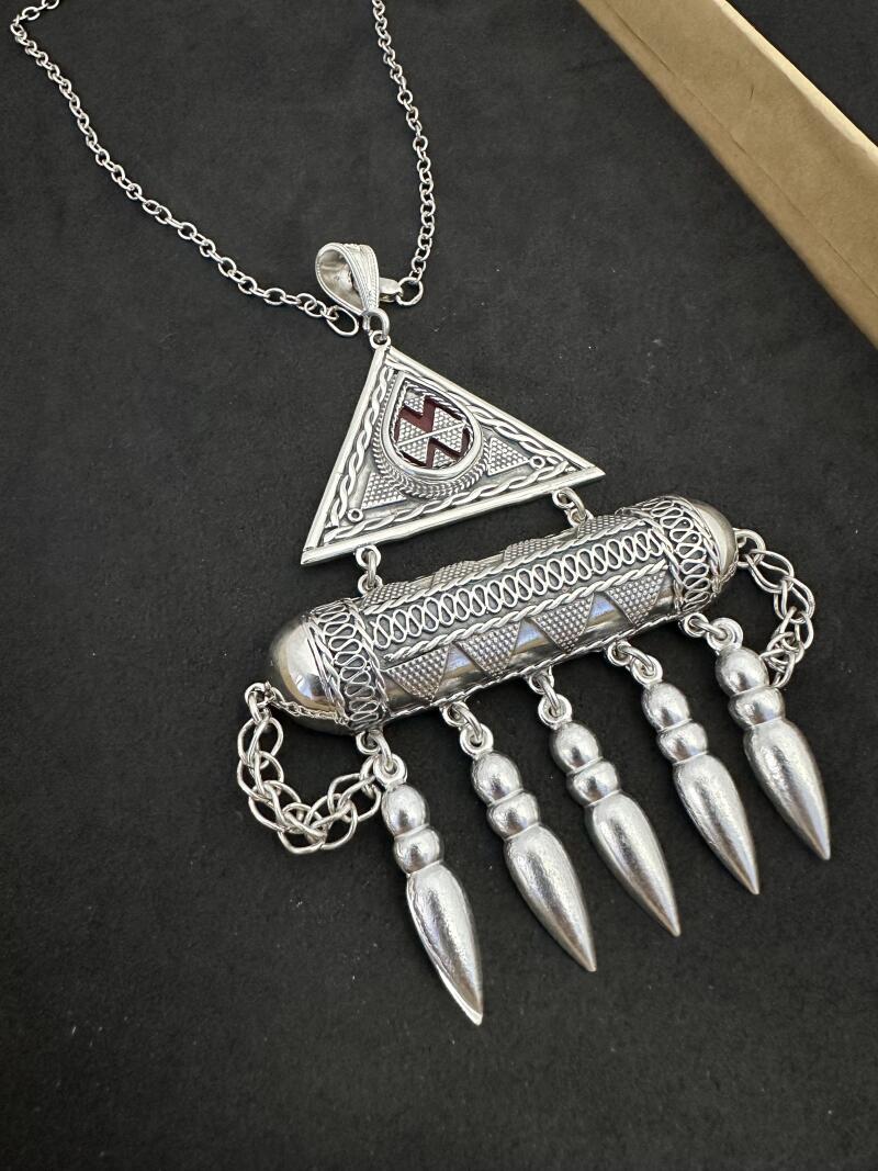 A silver necklace with a triangular pendant containing a red symbol, attached to a cylindrical piece with dangling silver elements, all on a silver chain.