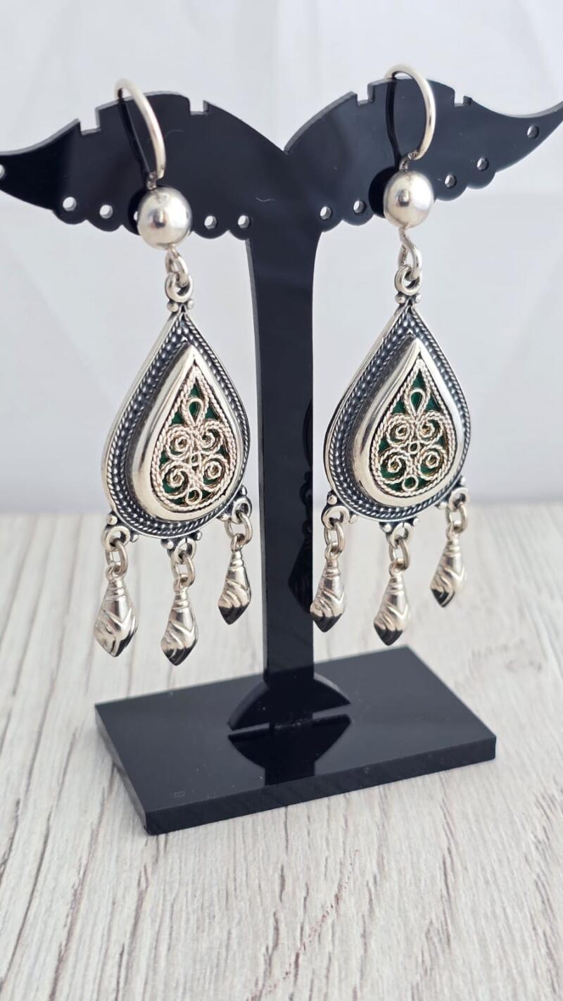 Two silver teardrop earrings with green filigree details hang from a black display stand.
