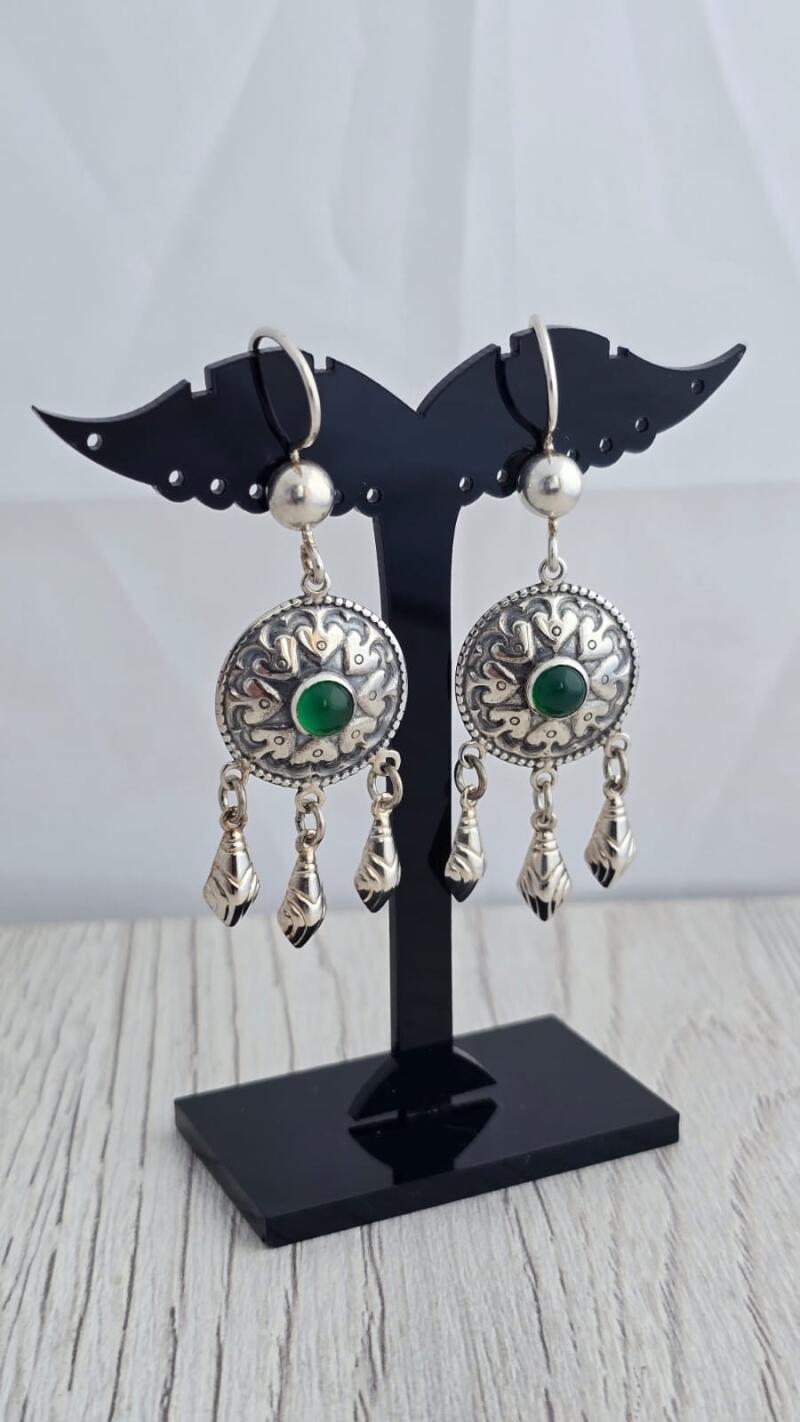 A pair of silver earrings with green stones and dangling ornaments are displayed on a black stand.