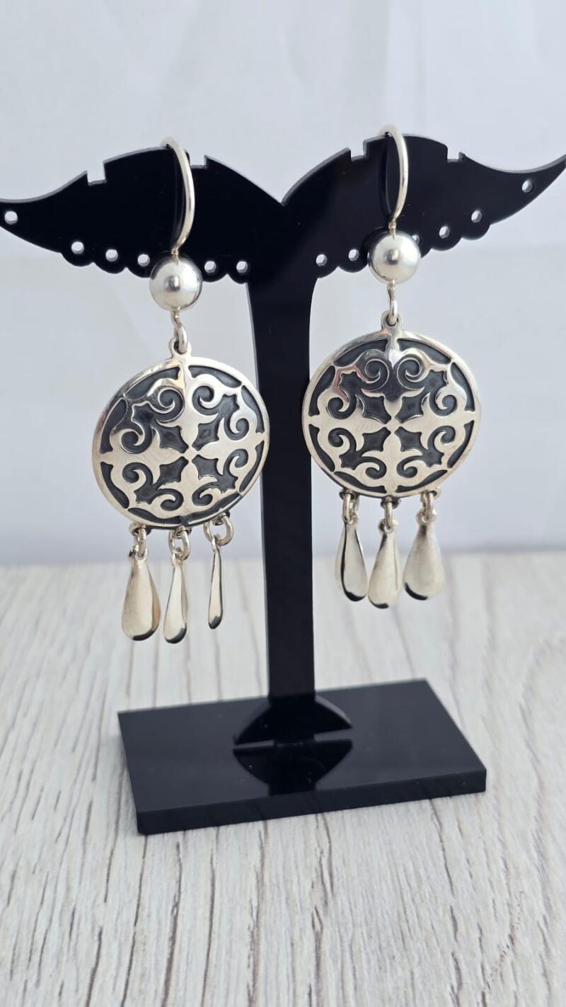 The image shows a pair of silver earrings with a circular design and teardrop pendants, displayed on a black stand.