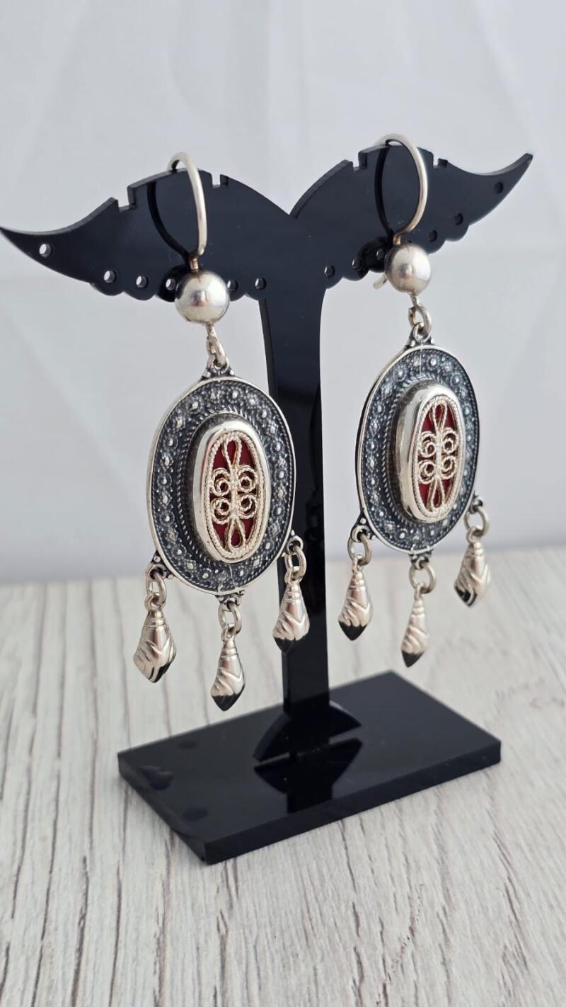 The image shows a pair of silver earrings with a red oval center, displayed on a black stand.