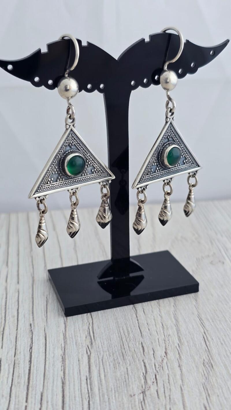 The photo shows a pair of silver earrings with green stones, hanging on a black display stand. The earrings are triangular in shape with three small charms hanging from each.