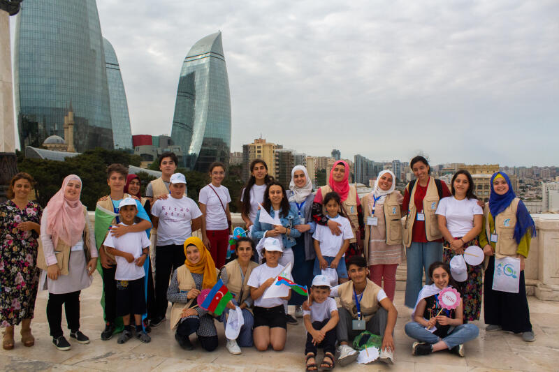 A group of young people, some holding Azerbaijani flags, poses for a photo with the Flame Towers in the background.