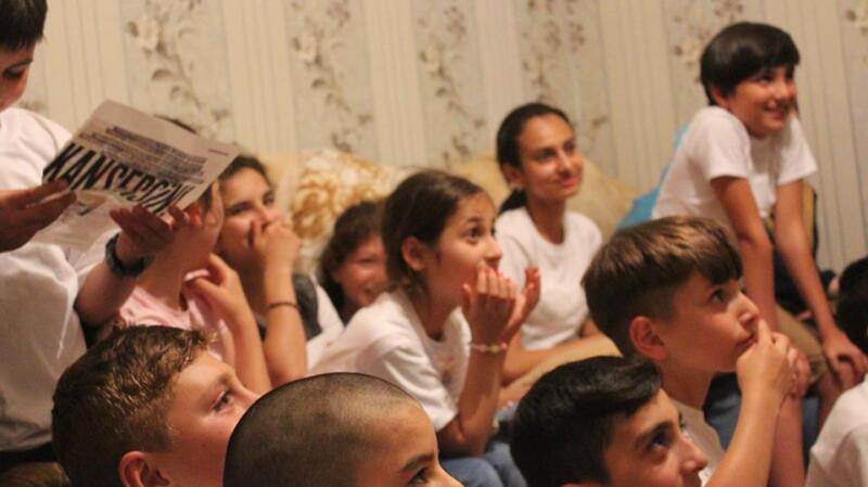 A group of children is sitting and looking upwards, with one child holding a newspaper that reads "KANSERCIN". The children are wearing white t-shirts and appear to be watching something with interest.