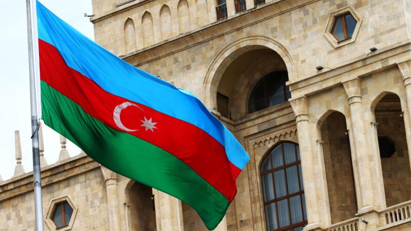 The Azerbaijani flag with a crescent and star is displayed in front of a stone building featuring arched windows and columns.