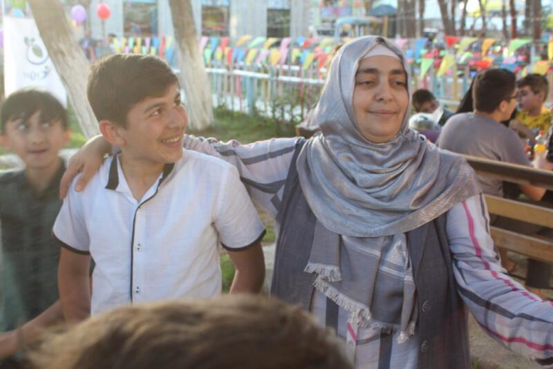 A woman in a hijab is smiling with her arm around a smiling boy, standing in front of a colorful outdoor setting.