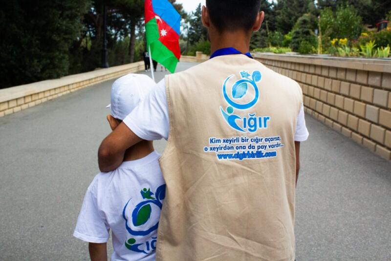 A person in a vest with the "Cığır" logo has their arm around a child wearing a matching shirt, with an Azerbaijani flag visible in the background. The vest also features the text "Kim xeyirli bir cığır açarsa, o xeyirdən ona da pay vardır.. www.cigirbirlik.com".