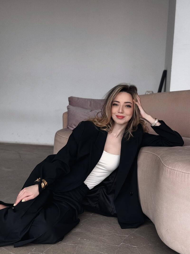 A woman with wavy blonde hair is sitting on the floor, wearing a black blazer over a white top and black pants, leaning against a light pink couch.