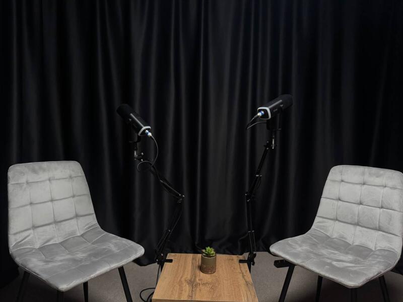 Two grey chairs face each other with microphones on stands between them, positioned on a small wooden table, against a black curtain backdrop.