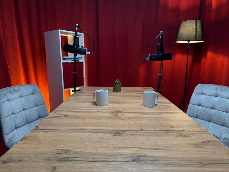 The photo shows a podcast studio setup with a wooden table, two mugs, microphones, chairs, a lamp, and a red curtain backdrop.