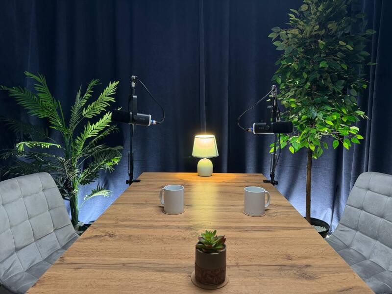 A podcast studio setup with a wooden table, two microphones, two mugs, plants, and a lamp against a dark blue curtain backdrop.