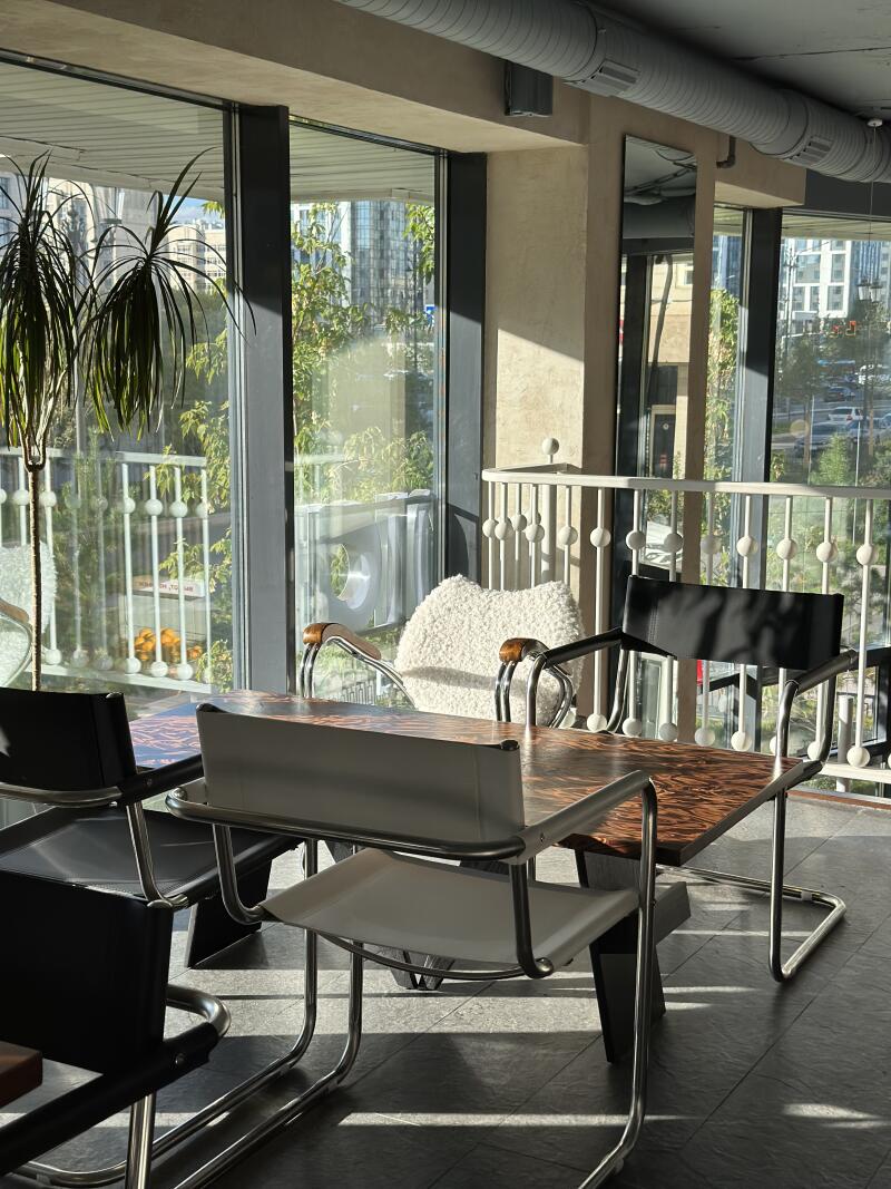 A brightly lit interior features a table surrounded by chairs of various designs, set against a backdrop of large windows offering a view of greenery and urban buildings.