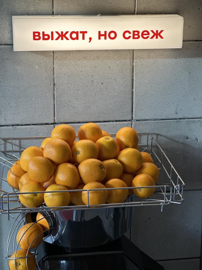 A wire basket is filled with oranges, positioned below a sign that reads "ВЫЖАТ, НО СВЕЖ" in red letters.