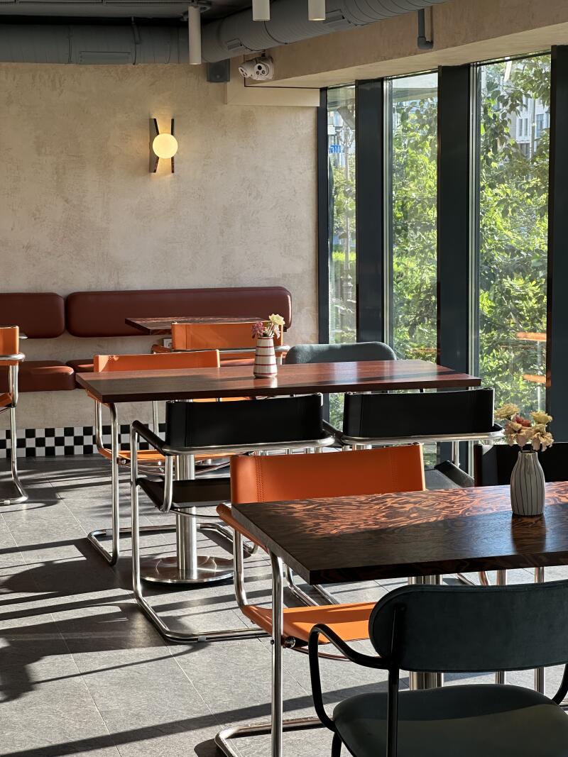 The interior of a café is shown with tables and chairs illuminated by natural light from a large window, with a brown cushioned booth along the wall and a checkered design at the base of the wall.