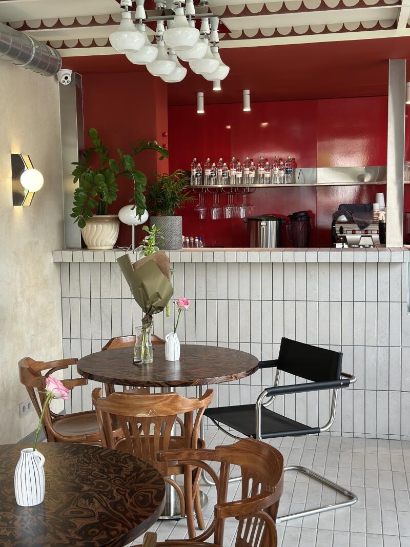 The photo shows a cafe interior with round wooden tables surrounded by wooden chairs and a single black chair, with plants and flowers as decorations. A red counter area is visible in the background, featuring a white tiled front and a shelf with bottles and glassware.