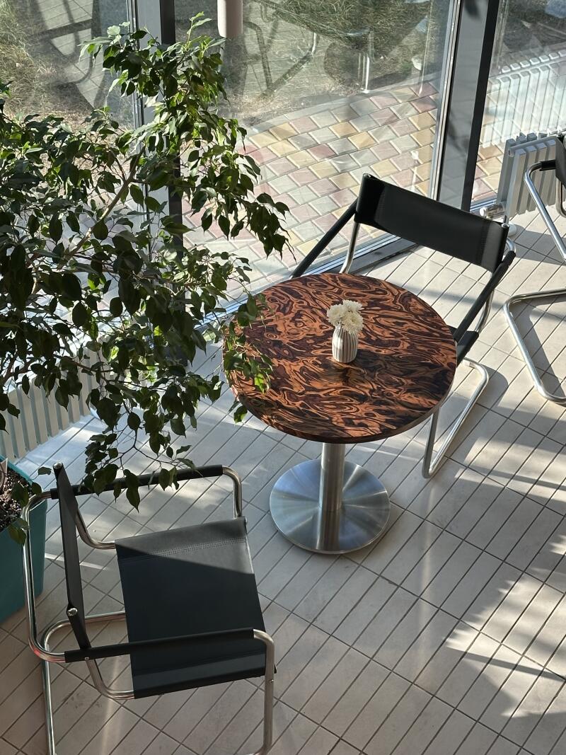 The image shows a round, patterned table with a vase of white flowers, surrounded by two chairs with black seats and silver frames, all set on a tiled floor near a window and a potted plant.