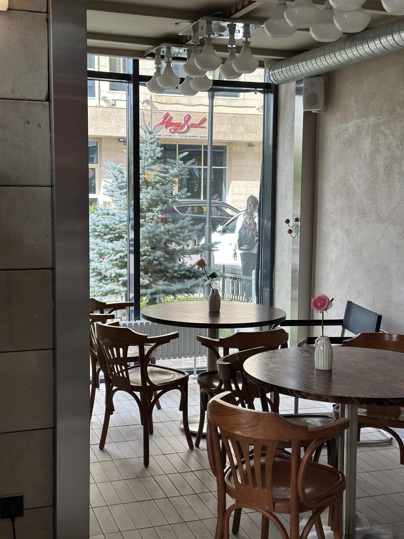 The interior of a cafe features wooden tables and chairs, each table adorned with a small vase holding a single rose; outside the window, views of the street include trees and building signage reading "Honey Soul" in cursive.