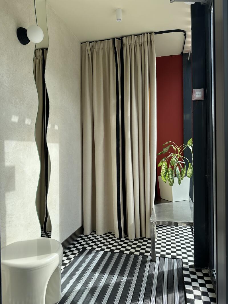 The image shows an interior with a distorted mirror, a stool, curtains with black stripes, and a plant, set against a black and white checkered and striped floor.