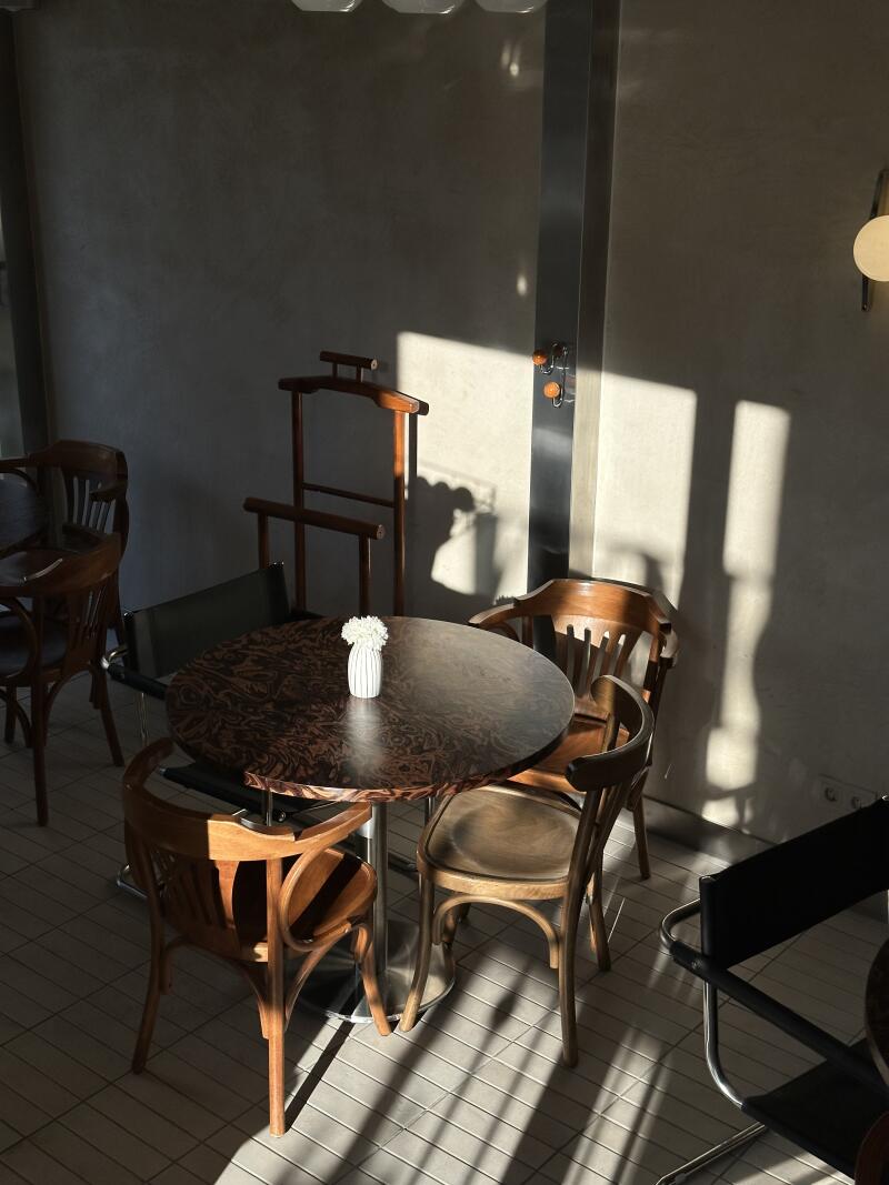 The photo shows a round, dark-wood table with a small white vase of flowers, surrounded by wooden chairs in a room with sunlight streaming through a window.