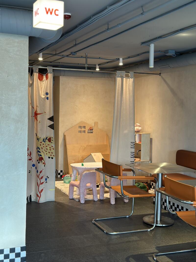 A room features a children's play area with a wooden house structure and small tables and chairs, along with standard seating at metal tables and a WC sign hanging from the ceiling.