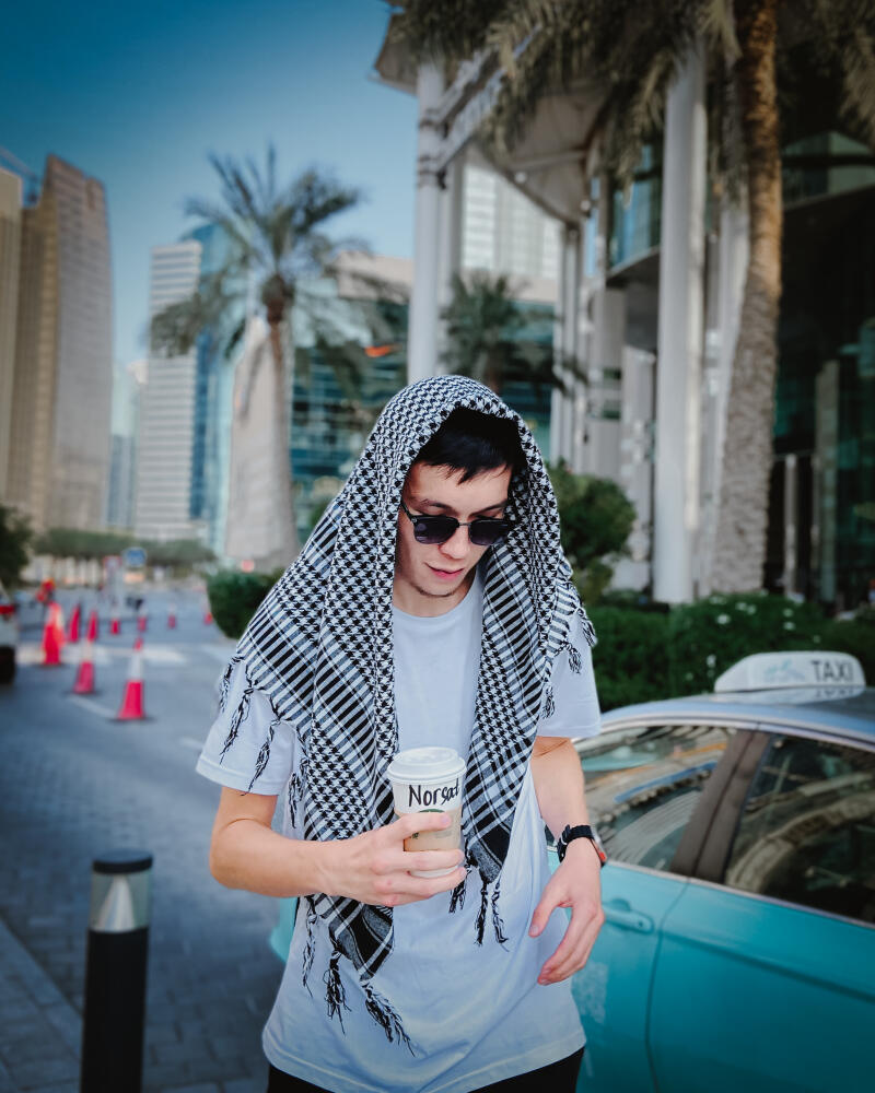 A person wearing sunglasses and a black and white keffiyeh is holding a coffee cup with the name "Norsad" written on it, standing on a city street with skyscrapers and palm trees in the background, next to a light blue taxi.