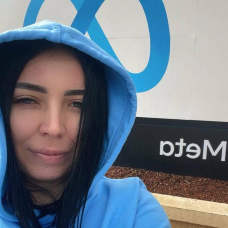 A woman with dark hair wearing a blue hoodie stands in front of a Meta logo.