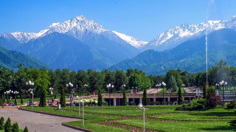 The landscape features a park with green trees and lawns, with a building and people visible in the background, set against a backdrop of majestic mountains with snowy peaks under a clear blue sky.