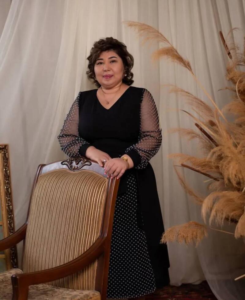 A woman with short brown hair is posing in a black polka-dot dress, leaning on an antique chair. She is wearing a necklace and a ring.