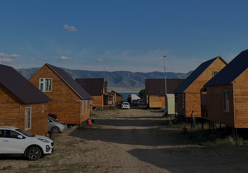 The image shows a row of wooden cabins on a sandy ground, with cars parked near some of them, and mountains and a body of water visible in the background under a blue sky.