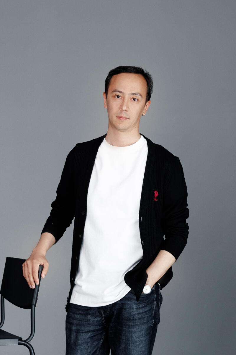 A man is standing with his hand on a chair, wearing a white t-shirt, a black cardigan with a red logo, and jeans, against a gray background.