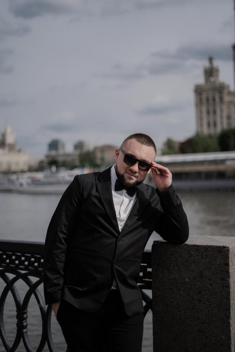 A man in a black suit and sunglasses leans against a stone railing near a river, with buildings in the background.