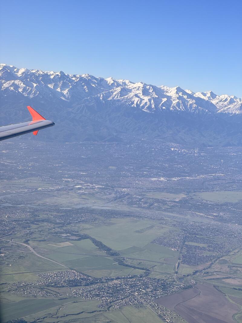 Aerial view featuring a landscape of fields and settlements leading up to a range of snow-capped mountains, with part of an airplane wing visible in the upper left corner.
