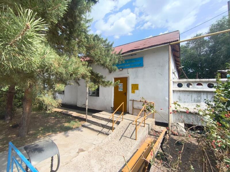 The image shows a one-story white building with a red roof and a yellow door, surrounded by greenery.