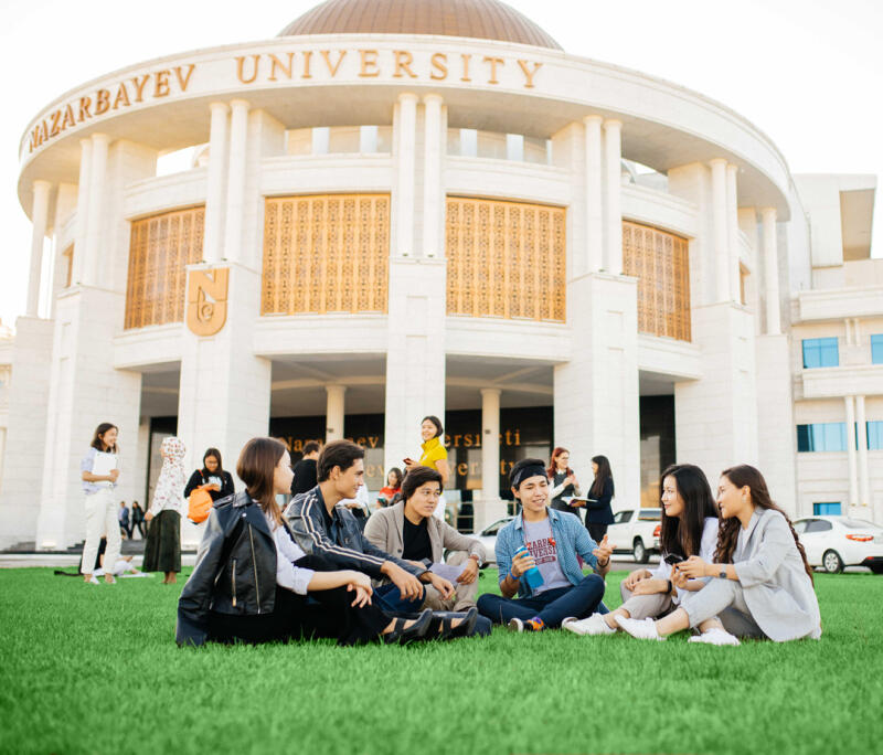 A group of students sit on the grass in front of Nazarbayev University, with the university's name displayed on the building's facade.