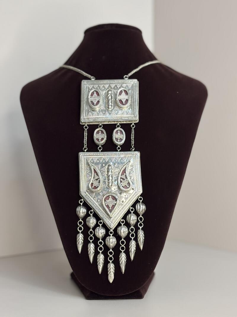 A silver necklace with a dark red accent is displayed on a brown velvet bust. The necklace has a chain and two pendants, with dangling silver beads and leaf-shaped charms.