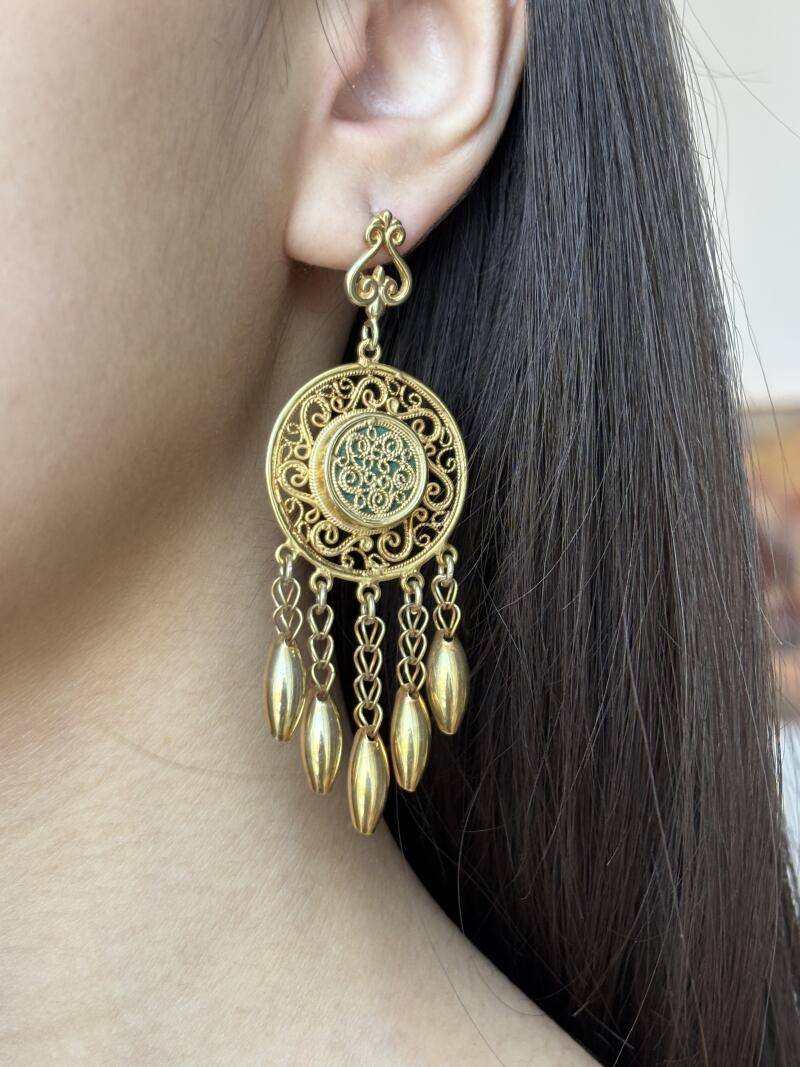A close-up shows a person's ear with a large, ornate gold earring featuring a circular design with dangling gold pendants.