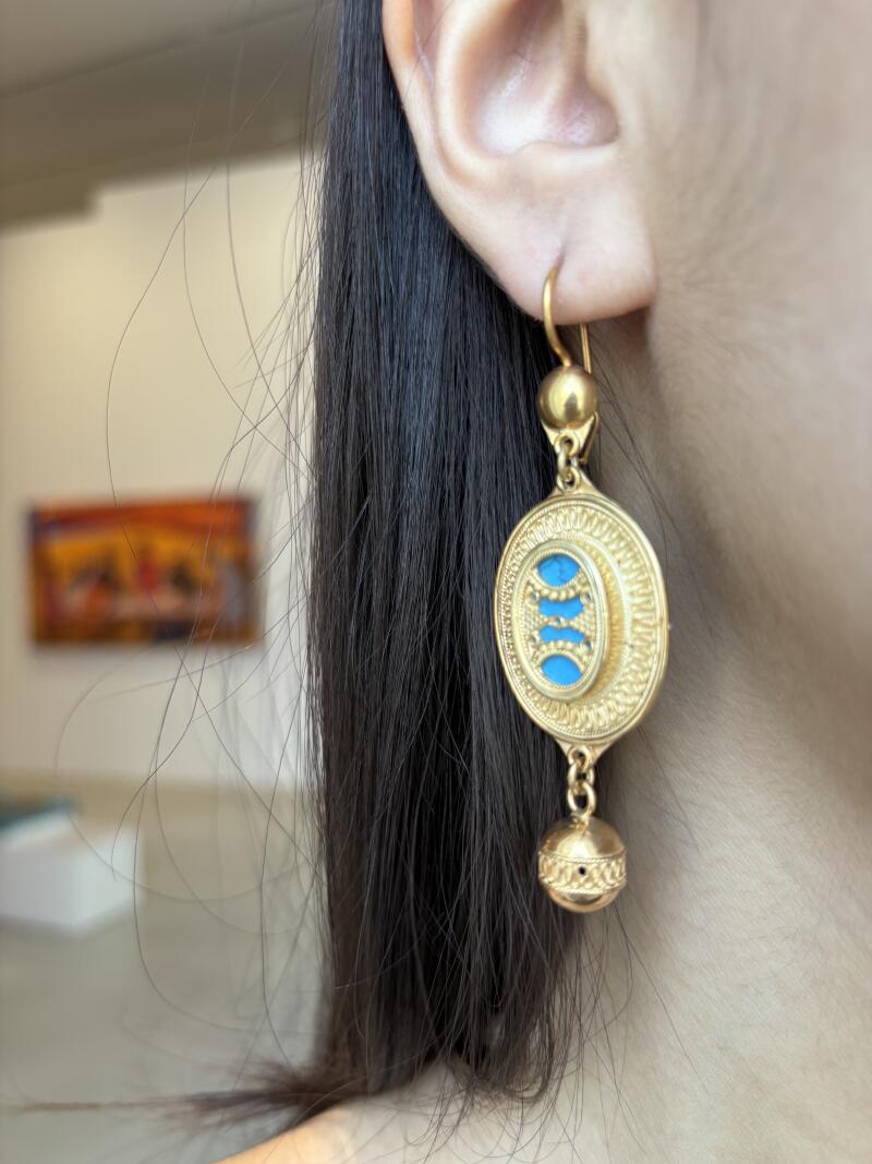 A person is wearing a gold earring with an oval turquoise inlay. The earring has a spherical accent at the top and bottom.