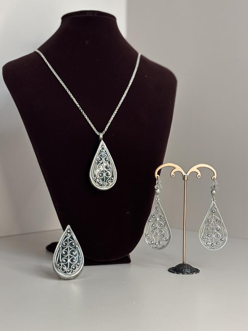 A jewelry set is displayed, including a teardrop pendant necklace on a brown display, a teardrop ring, and teardrop earrings on a gold stand, all featuring silver filigree.
