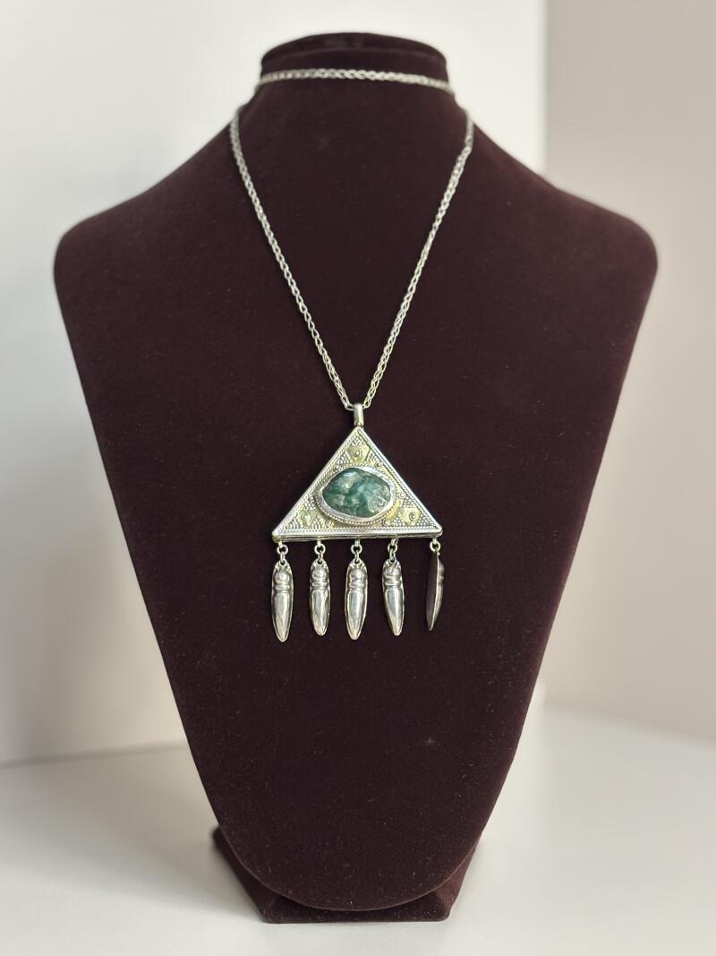 A silver chain necklace with a triangular pendant featuring a green stone and dangling silver charms is displayed on a brown velvet mannequin.