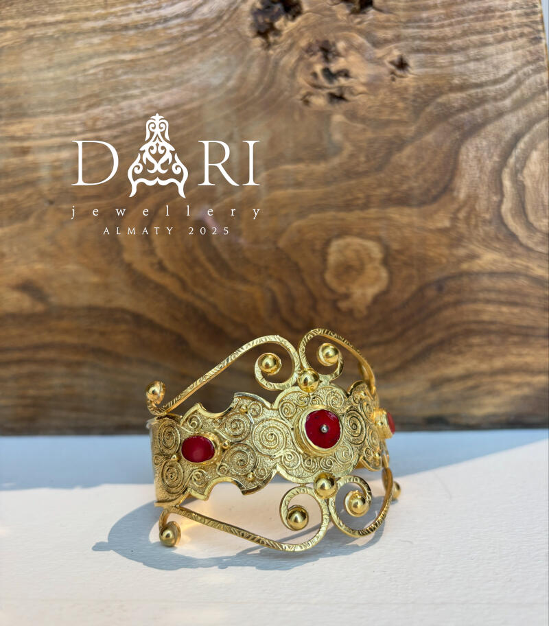 A gold-colored, ornate bracelet with red stones is displayed against a wood grain background, with the text "DARI jewellery ALMATY 2025" visible above it.