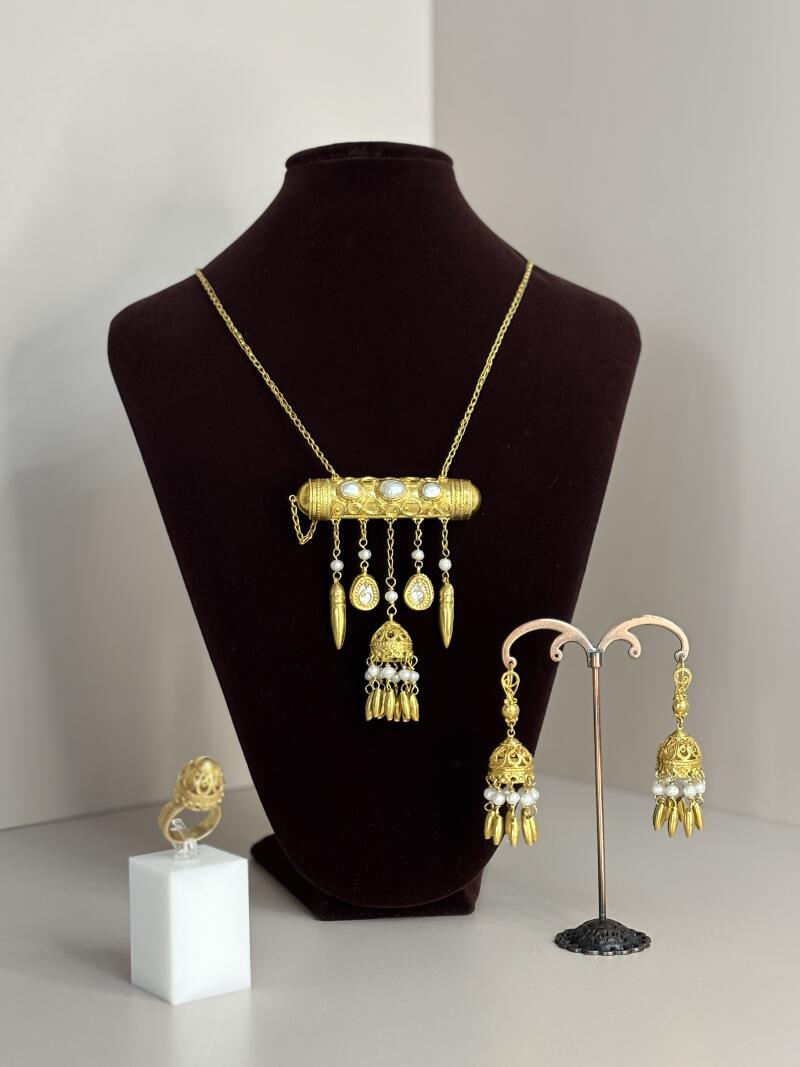 The image shows a set of gold-colored jewelry consisting of a necklace with a cylindrical pendant, a ring, and earrings with pearl accents, displayed against a brown velvet bust and a white surface.