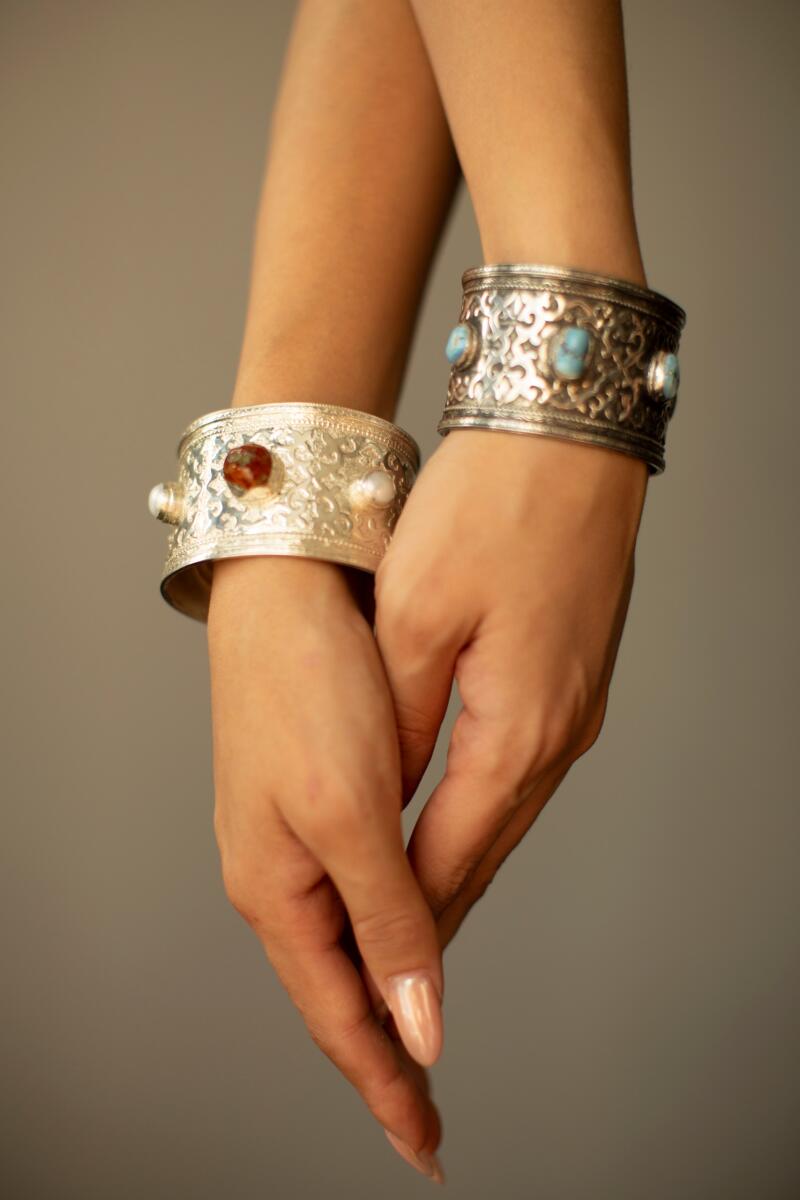 The image shows two hands adorned with ornate silver bracelets, one featuring an amber stone and pearls, and the other decorated with turquoise stones.