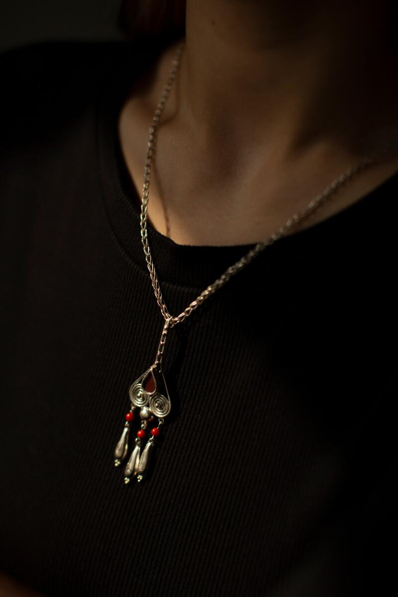 A person wears a silver necklace with a pendant featuring red accents and dangling silver and red beads, set against a black shirt.