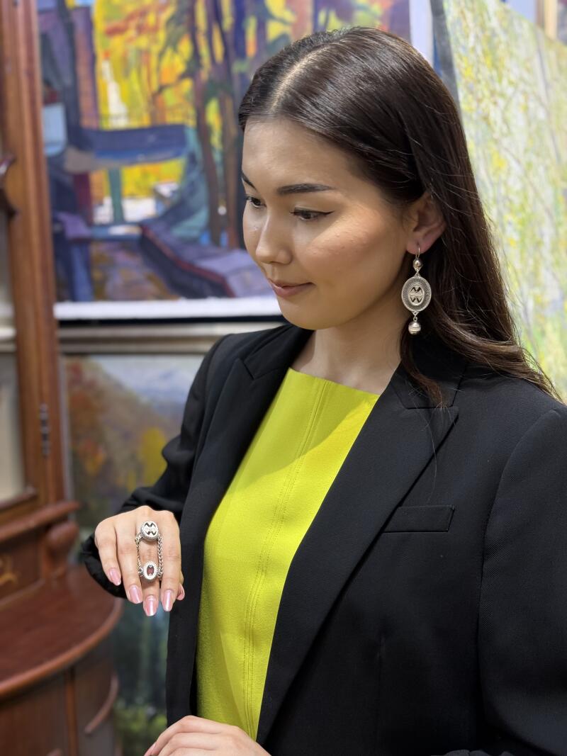 A woman in a yellow shirt and black blazer is wearing a ring and earrings with a Kazakh national pattern.
