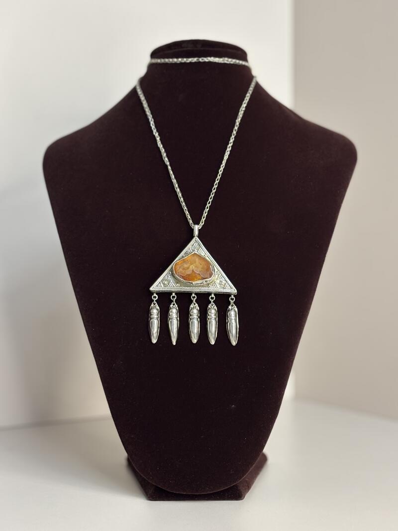 A silver necklace with a triangular pendant featuring an amber stone and dangling silver charms is displayed on a dark brown mannequin.