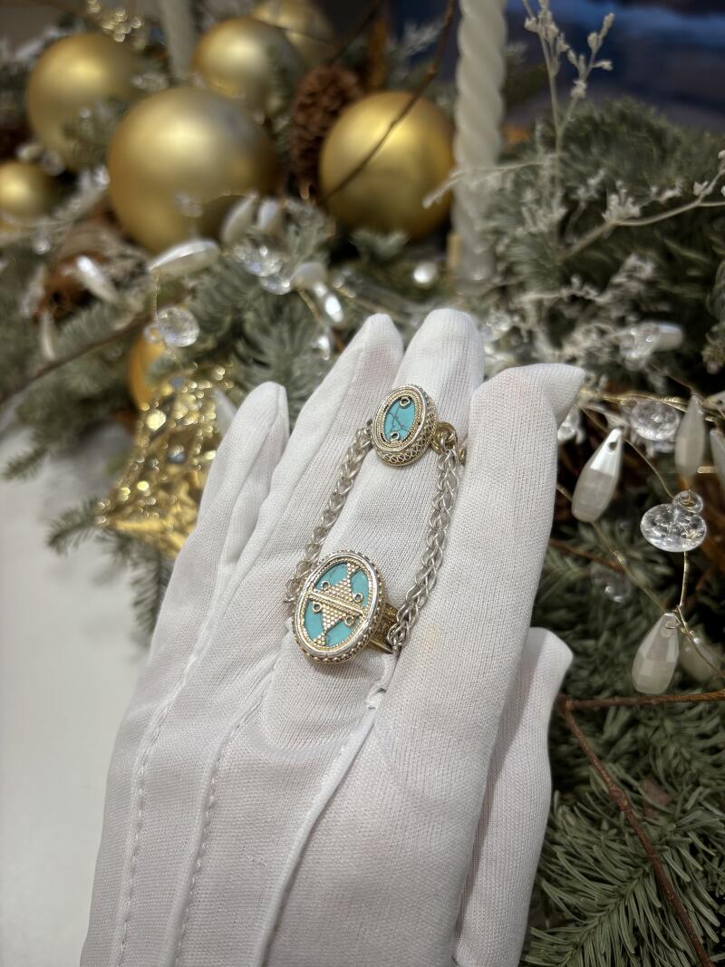 A hand in a white glove displays a silver chain ring with turquoise oval stones, set against a background of gold ornaments and greenery.