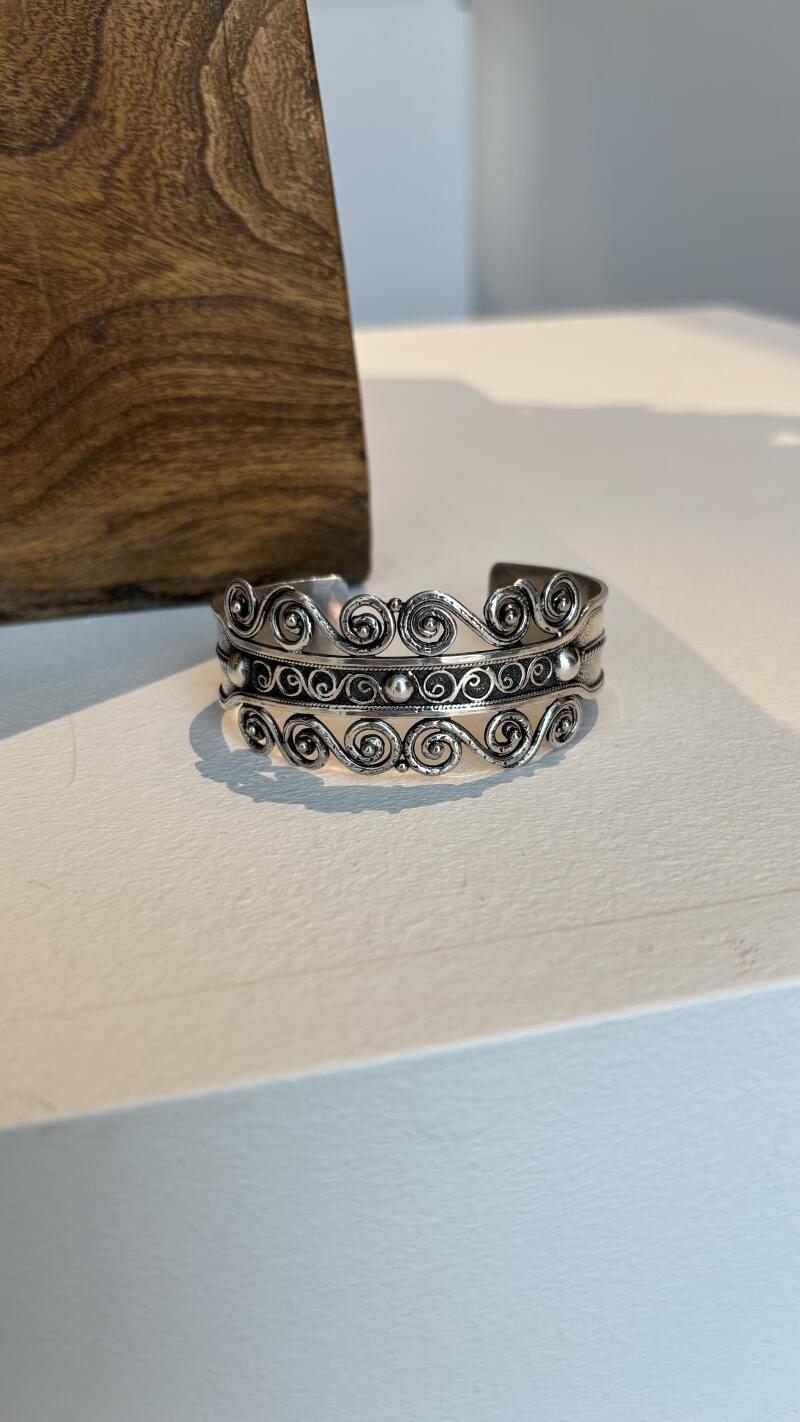 The image shows a silver bracelet with intricate swirl designs on a white surface, next to a wooden object.