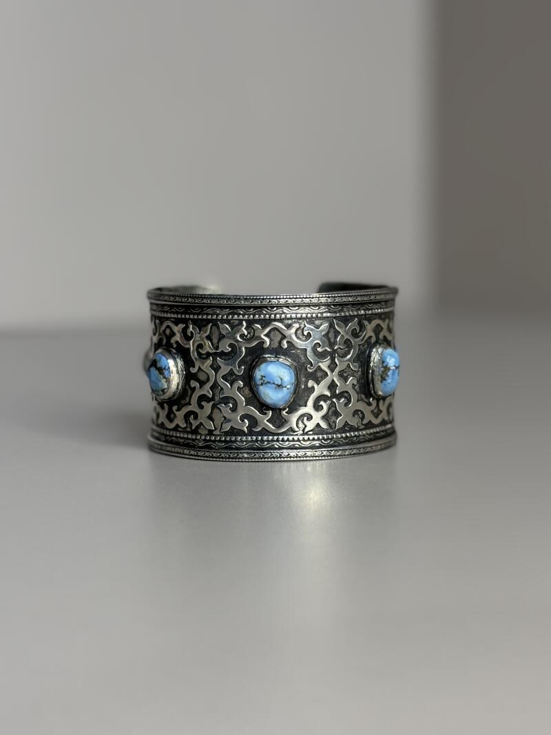 A silver cuff bracelet with ornate patterns and three light blue stones is displayed.