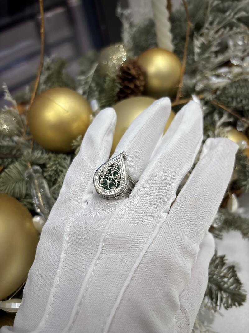 A hand in a white glove presents a silver ring with a green, decorative stone, against a backdrop of gold ornaments and pine branches.