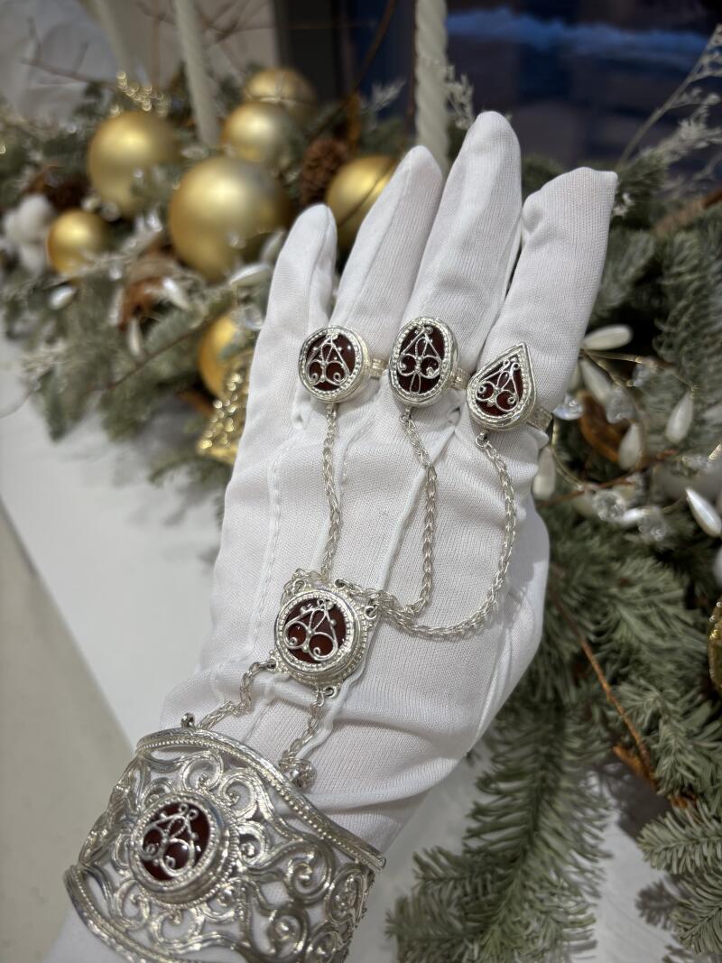 A white-gloved hand models a silver hand chain with red stone insets, featuring a wide cuff and rings connected by delicate chains, set against a background of holiday greenery and golden ornaments.